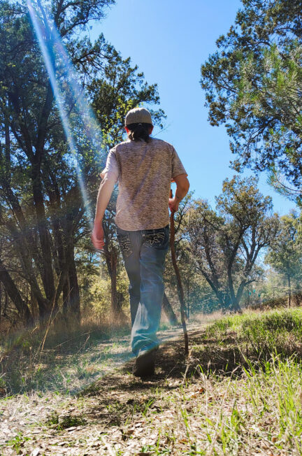 a slim man walking in jeans and white tee shirt with walking stick lens flare sunbeam shining down in peaceful forest path
